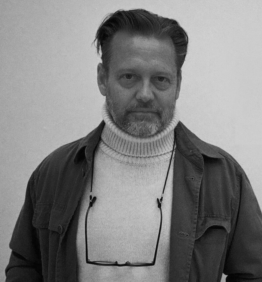 A black and white, head and shoulders portrait of a man, Martin Stigsgaard, wearing a turtleneck sweater and an open jacket, with glasses hanging from his neck.