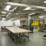 Interior view of the fabrication shop at the affordable Spitzer School of Architecture, featuring a communal work desk on the left with various power tools on the right.