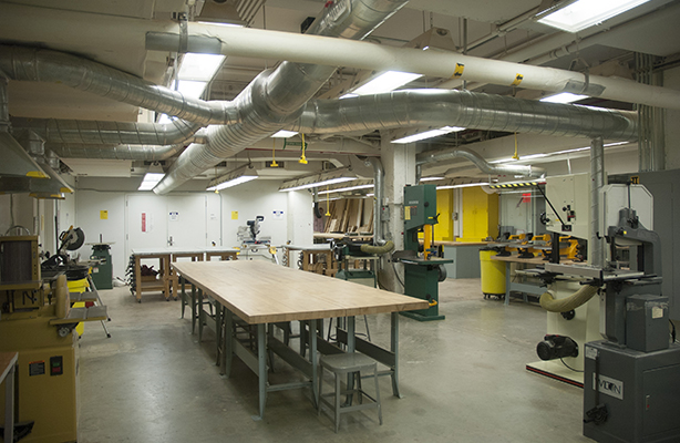 Interior view of the fabrication shop at the affordable Spitzer School of Architecture, featuring a communal work desk on the left with various power tools on the right.