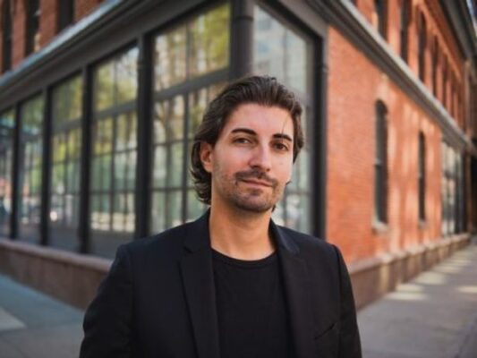 Alex Nizhikhovskiy, dressed in a black blazer and shirt, looks at the camera.