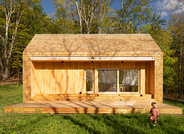A rectangular cabin entirely clad in light wood shingles on its roof and side walls, sitting in a grassy clearing surrounded by trees. The front features a deep, recessed facade of vertical wood planks, large glass sliding doors, and a wide wooden deck spanning the base. Tree shadows fall across the structure, and a small child in pink clothing is walking in the grass in the lower right foreground.