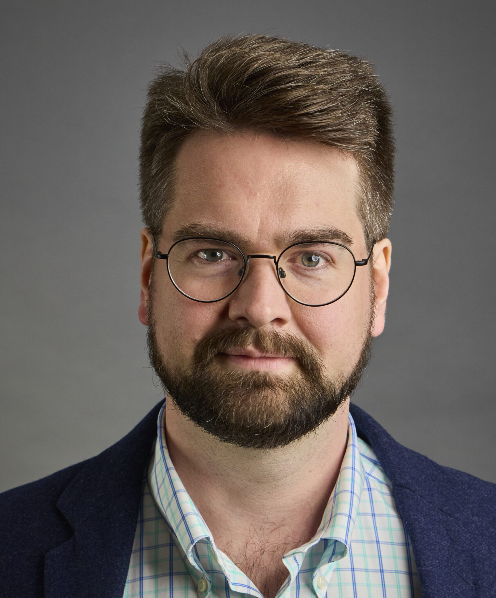 A man with brown hair and a full beard, wearing round wire-rimmed glasses, a blue and white plaid button-down shirt, and a dark blue blazer. He is looking directly at the camera with a neutral expression against a plain gray background.