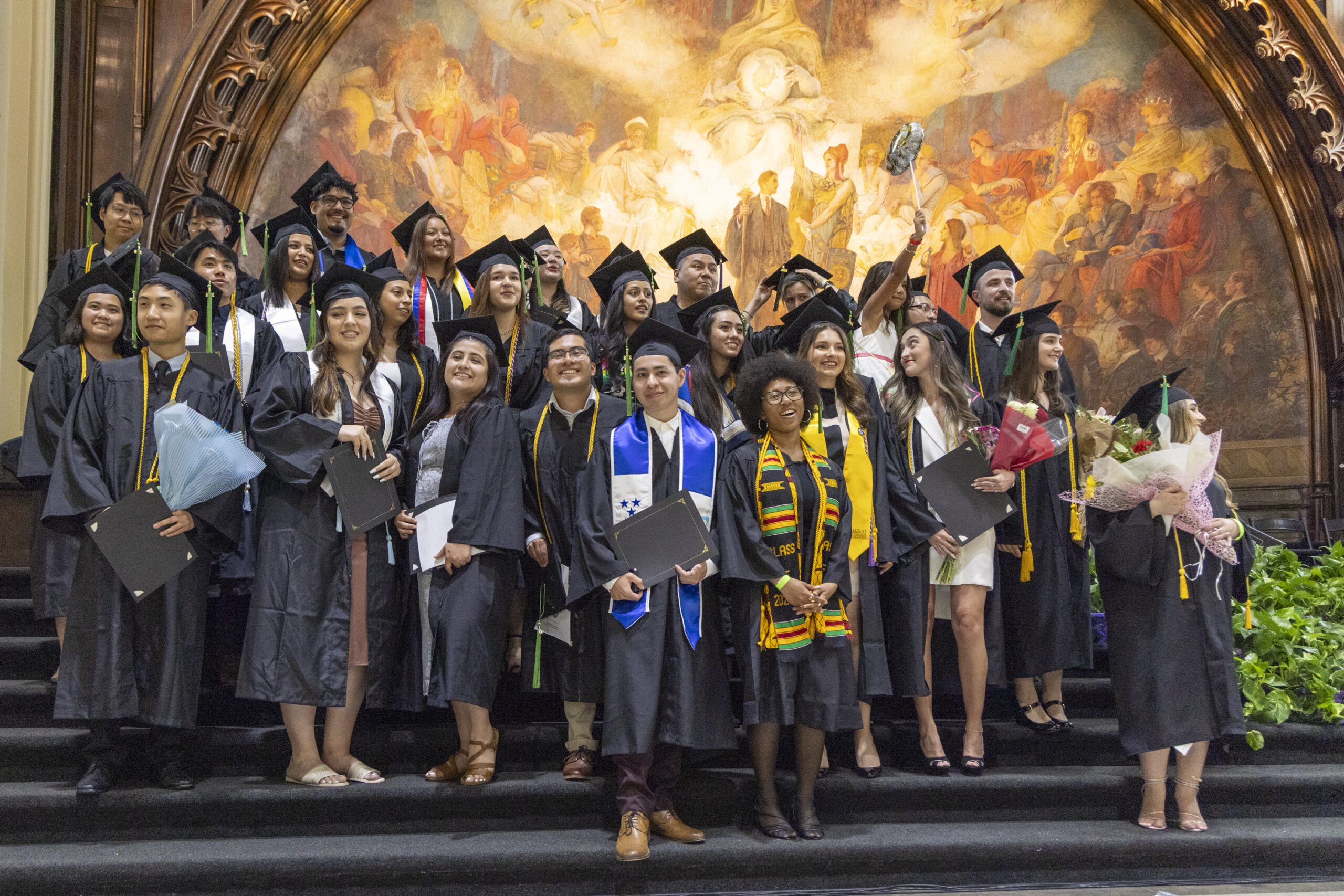 Graduating students in their commencement regalia stand on the platform.
