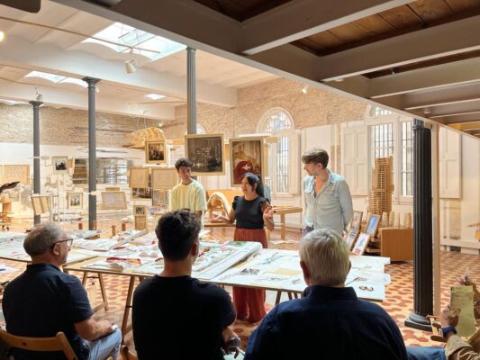 Three students stand in front of their architectural project and present it to a group of seated reviewers.
