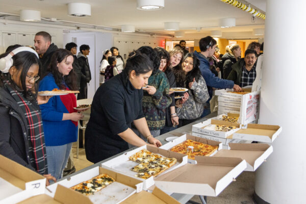 Students stand in front of a table of pizzas in open cardboard boxes. Students are holding plates with pizza.