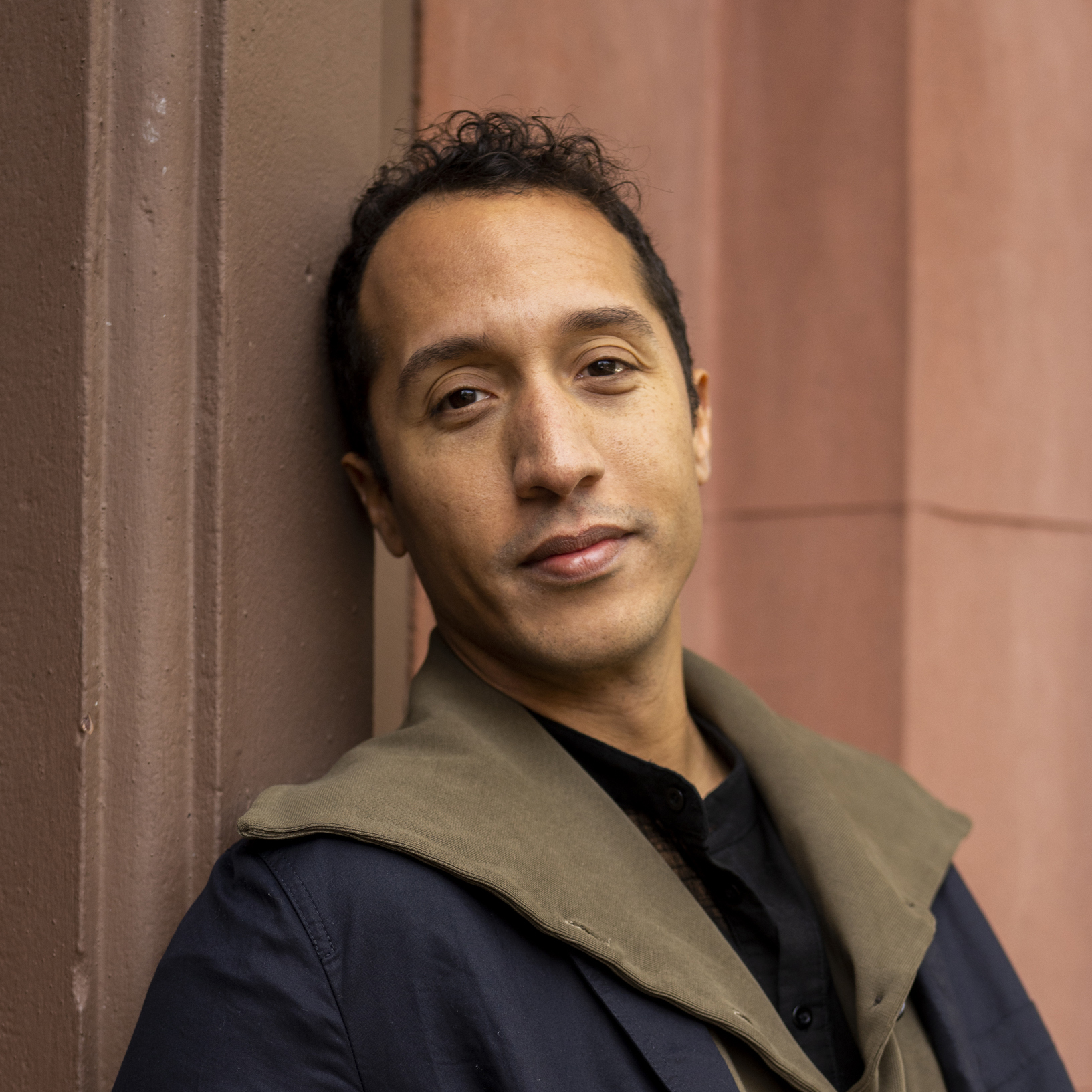A head-and-shoulders portrait of Jerome Haferd, a man with short dark curly hair, looking directly at the camera. He is wearing a dark jacket with a tan or olive-colored collar, and a dark shirt underneath. He is positioned against a textured brown wall, which appears to be part of a building.