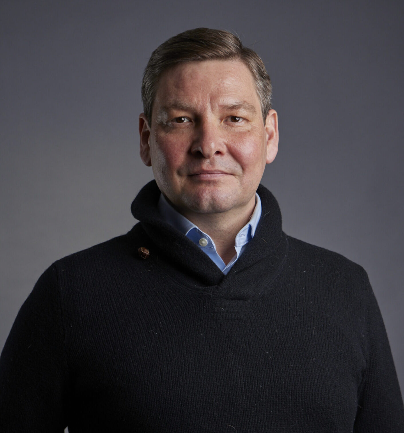 A portrait of a man with short brown hair, wearing a black shawl-neck sweater over a light blue collared shirt. He is looking directly at the camera with a neutral expression and has his right hand casually placed in his pocket. The background is a solid, dark gray.