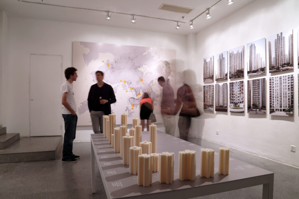The image shows the interior of an art gallery. Several people are viewing the exhibits. On the left, two figures are looking at a large map on the wall. In the center, a person is standing in front of a table displaying a series of small, cylindrical architectural models. To the right, a set of framed photographs of building exteriors are hung on the wall. The gallery has white walls and a gray floor.