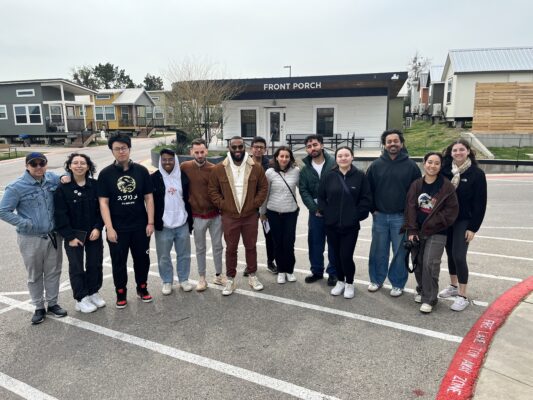 A group of 14 adults and young adults stands in a line outdoors, smiling at the camera. They are diverse in appearance, with varied skin tones and hair colors. The building behind them reads 