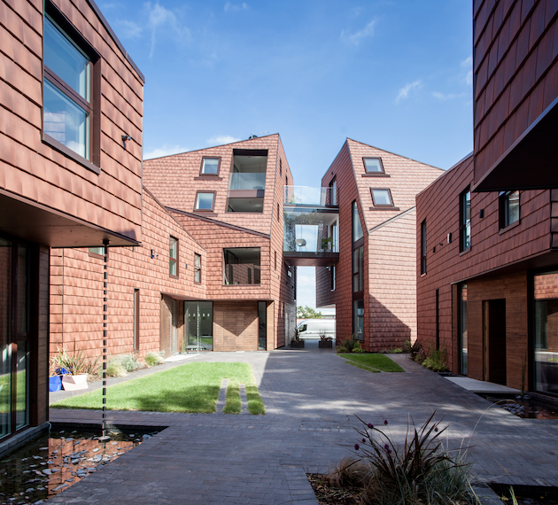 The image shows an exterior view of Kaolin Court, designed by Baca Architects Ltd and Stolon Studio. The buildings feature a distinctive reddish-brown shingle siding and angled rooflines. A central walkway leads through the complex, with a glass-enclosed bridge connecting two of the structures. A small pond and some greenery are visible in the foreground.