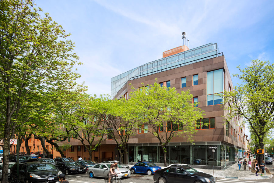 An exterior shot captures the Brooklyn Children's Museum, located in New York, United States. The building features a modern design with a mix of brown and copper-toned cladding, large windows with orange frames, and a glass-enclosed upper level. Lush green trees line the sidewalk in front of the museum, partially obscuring the view at street level. Several parked cars and a few pedestrians are visible on the street, suggesting a typical urban scene on a day with a partly cloudy sky.
