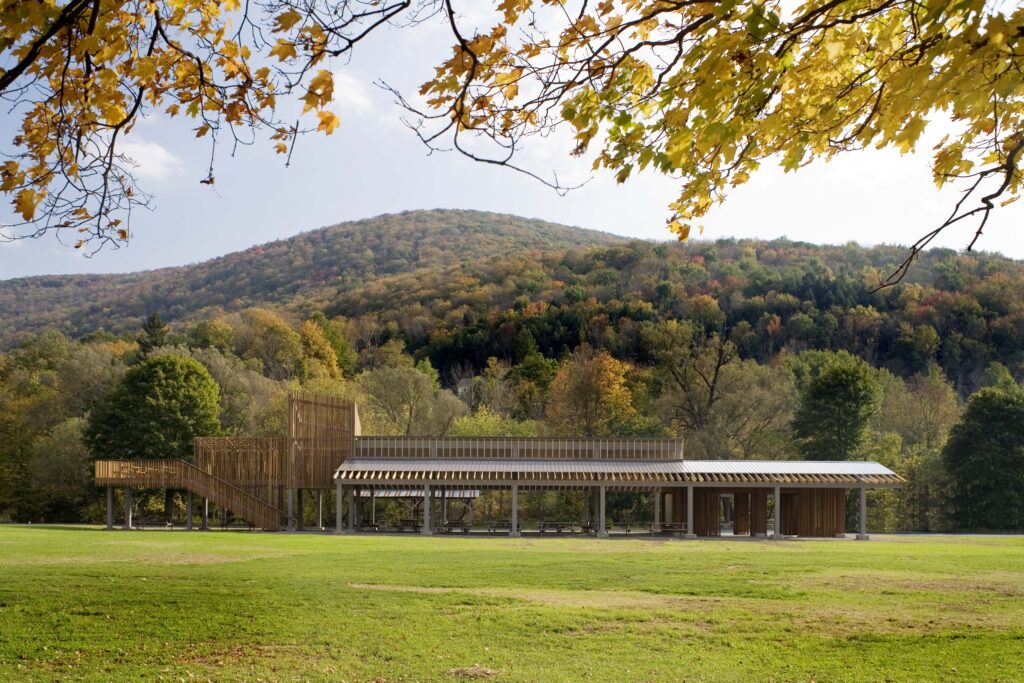 The image shows the Margaretville Pavilion, with a backdrop of a hill covered in fall foliage. The pavilion is a wooden structure with a long, open-sided roof. There's a set of steps leading up to a platform on the left side of the pavilion. The scene is set in a grassy field with trees surrounding the area.