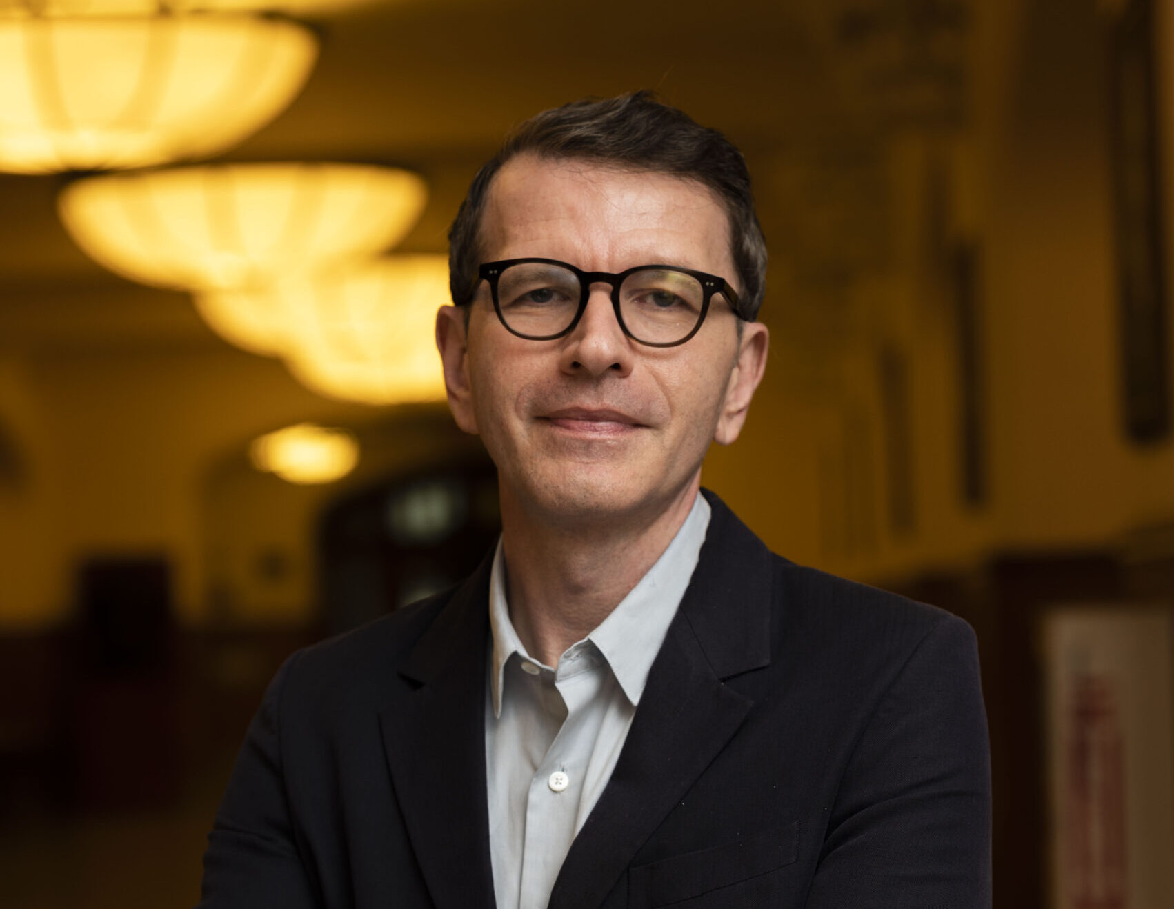 A light-skinned man with close-cropped dark hair and dark-rimmed glasses stands with his arms crossed and a slight smile. He is wearing a dark suit jacket over a light-colored collared shirt, unbuttoned at the neck. He is positioned from the waist up, facing forward. The background is a dimly lit hallway with a series of overhead lights creating a warm glow.