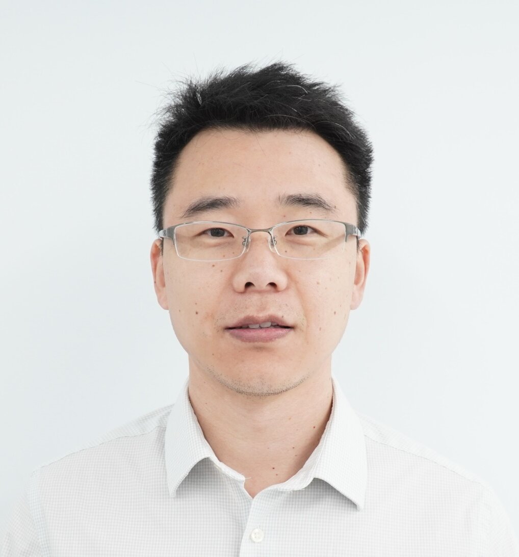 A headshot of a man with short black hair and glasses, looking directly at the camera with a neutral expression. He is wearing a light-colored, collared shirt. The background is solid white.
