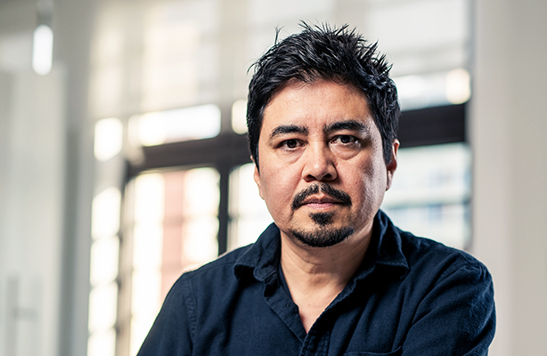 A professional headshot features Frank Melendez, a man with a dark complexion and dark hair, directly facing the camera with a serious expression. He has short, dark hair that is slightly graying at the temples, and a well-maintained dark goatee and mustache. He is wearing a dark, collared shirt. The background is softly blurred, showing an indoor setting with hints of windows and natural light.