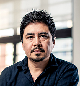 A professional headshot features Frank Melendez, a man with a dark complexion and dark hair, directly facing the camera with a serious expression. He has short, dark hair that is slightly graying at the temples, and a well-maintained dark goatee and mustache. He is wearing a dark, collared shirt. The background is softly blurred, showing an indoor setting with hints of windows and natural light.