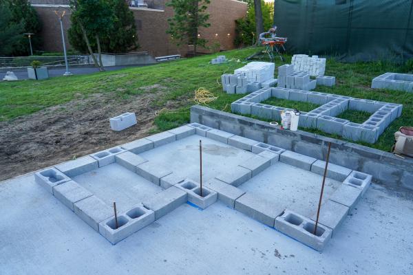 A construction site at dusk shows the initial stages of a design-build project. In the foreground, a foundation of concrete blocks is laid out in a zig-zag pattern on a concrete slab, with rebar protruding vertically from some of the blocks. Behind this, more concrete blocks are stacked, some forming small square frames, and building materials like buckets are visible. In the background, a grassy hill slopes upwards towards some trees and a building, and a green construction tarp covers a structure to the far right. The overall impression is one of active, ongoing construction.