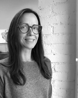 A black and white portrait shows a woman with dark, shoulder-length hair and large, dark-rimmed glasses smiling slightly. She is wearing a light-colored, possibly knit, long-sleeved top. The background features a textured white brick wall on the right and a softly blurred, lighter area on the left.