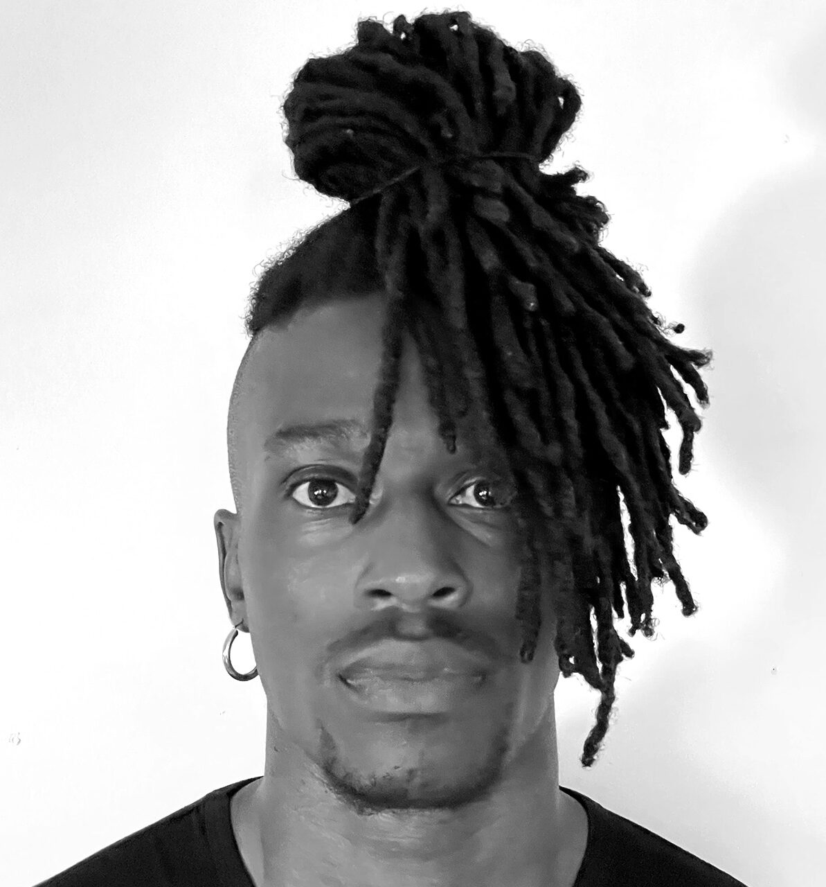 A black and white close-up of a man looking directly at the camera. He has dark skin, a light mustache and soul patch, and wide-set eyes. His hair is styled in long dreadlocks, with the majority pulled up into a high bun on top of his head, leaving some dreadlocks hanging down past his left eye. The sides of his head are closely shaved, and he wears a single large hoop earring in his left ear. He is wearing a dark, possibly black, t-shirt. The background is a plain white wall.