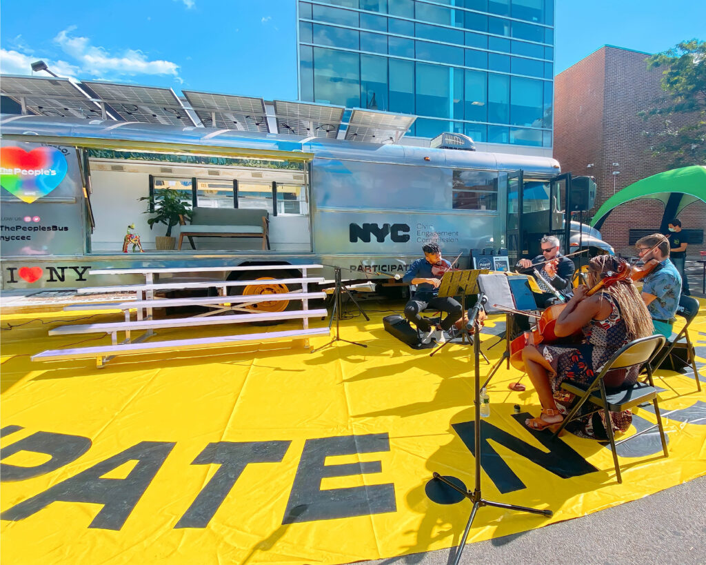 A shiny, silver bus with solar panels on its roof is parked on a bright yellow mat. The side of the bus is open, revealing a minimalist interior with a bench. Four musicians are playing string instruments on the mat in front of the bus. A tall, modern glass building is visible in the background.