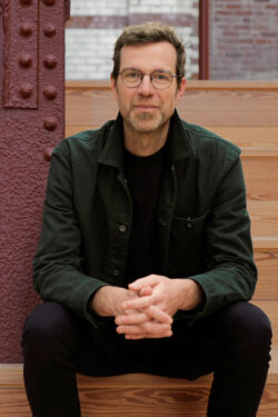 A man with glasses and a beard is sitting on wooden steps. He is wearing a dark green jacket over a black shirt and black pants, with his hands clasped together in his lap. The background includes a reddish-brown metal column and a brick wall.