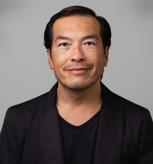 A man with short, dark hair and a mustache is smiling slightly at the camera. He is wearing a black jacket over a black v-neck shirt against a gray background.