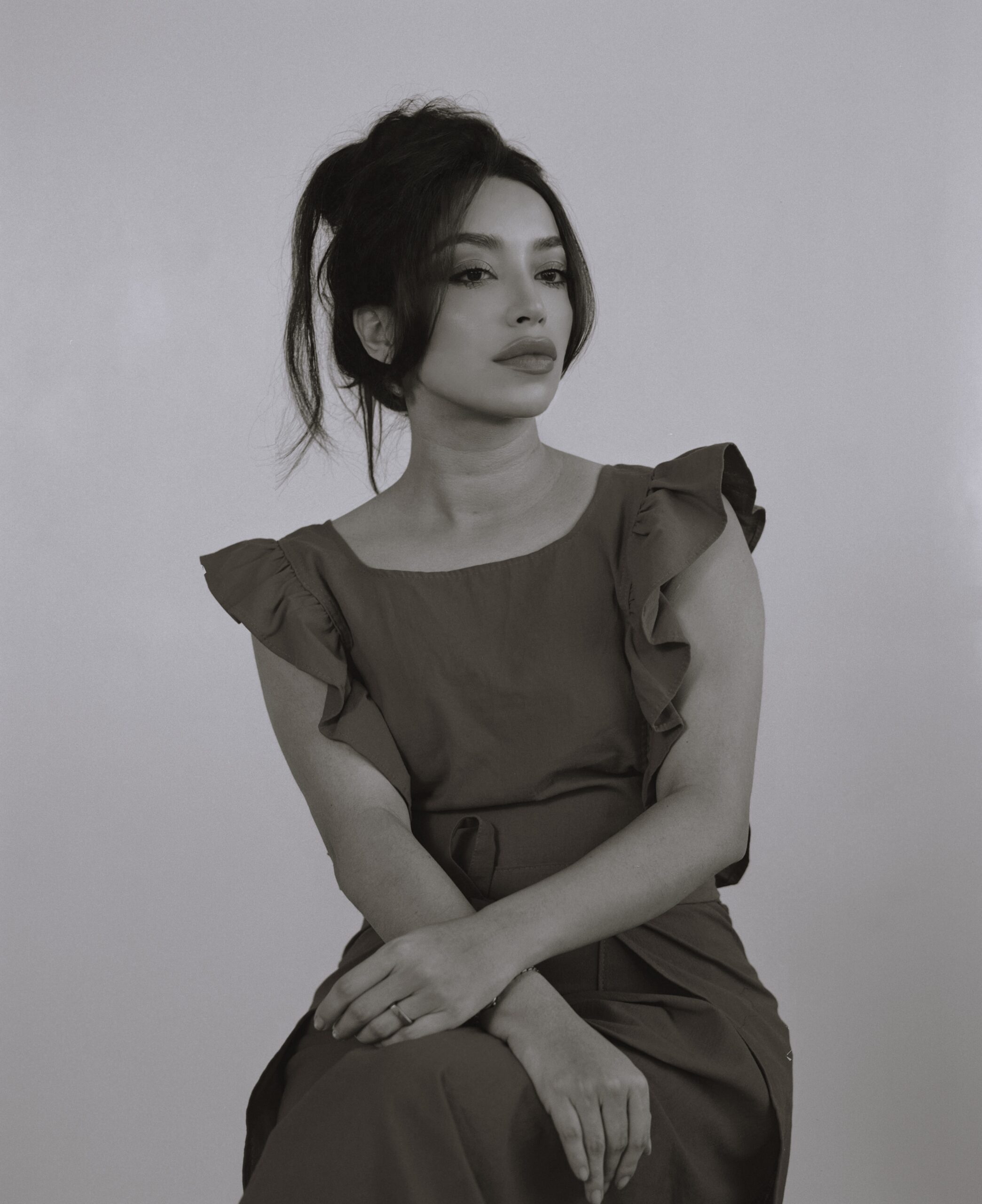 A black and white studio portrait of a woman sitting and looking off to the side. She has her hair in a messy bun, with loose strands framing her face, and is wearing a top with ruffled sleeves. Her arms are crossed, and she is wearing a ring on her left hand.