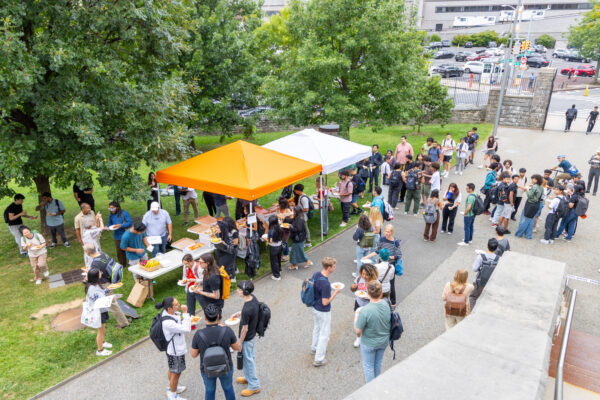 A crowd of people, including many students, stands around tables under two pop-up tents—one orange and one white—outdoors on a grassy area. They are getting and eating food, with some holding paper plates. A paved walkway runs through the scene, and a few people are walking or standing along it. In the background, there are more trees, a parking lot, and a city building.