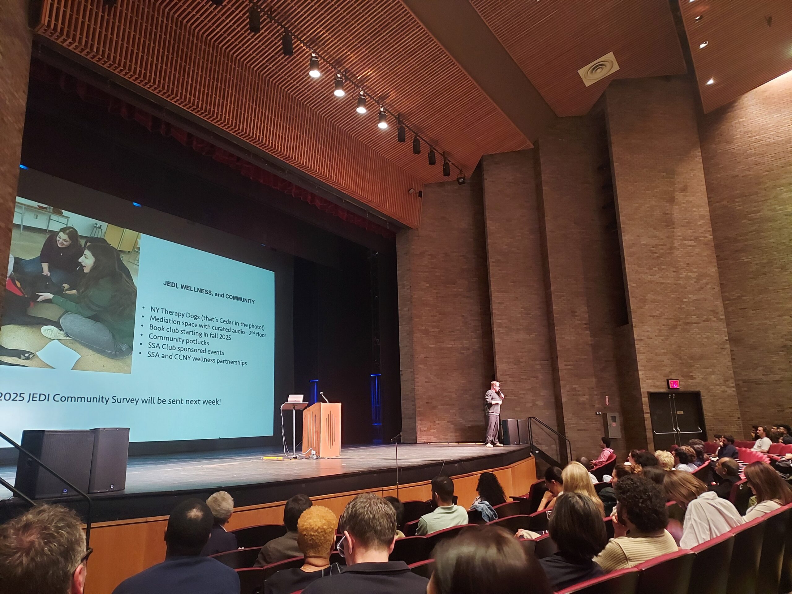 A man stands on a stage in a large auditorium, giving a presentation to an audience. He is on the right side of the stage, next to a wooden podium. A large projection screen behind him displays a presentation slide with the title 