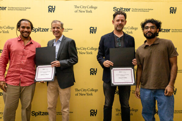 Against a bright yellow backdrop with university branding, two pairs of men are shown, each pair holding an academic award. All four men are smiling, two are casually dressed and two are in blazers.