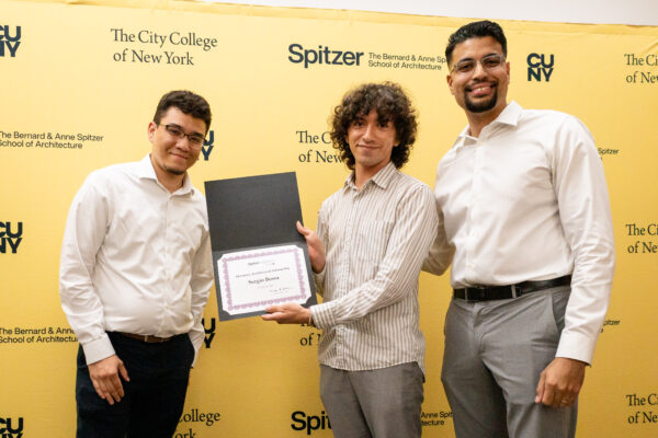 Three smiling men in business casual attire are holding an award certificate against a yellow university-branded background.