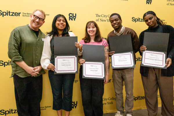 A group of five people, including an instructor or official and four students, stands against a yellow university wall while holding four separate academic awards.