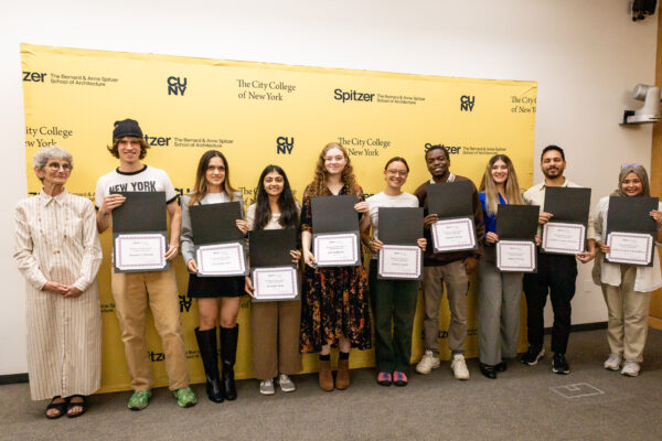 A large group of nine people is pictured holding eight academic awards at a ceremony, all standing before a yellow wall with university logos.