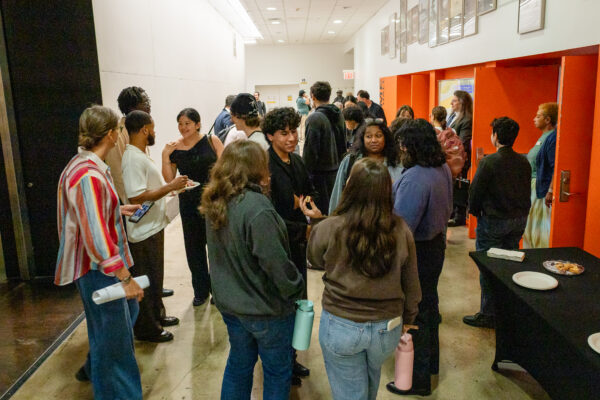 A crowd of people is mingling and socializing in a brightly lit hallway or reception area. The group is diverse, with several people standing in small circles, while others walk in the background. The area features a black table with plates of food and bright orange walls contrasting with a darker floor.