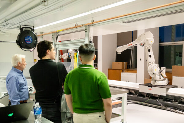 Three people stand with their backs to the camera, observing a robotic arm in a university robotics or fabrication lab.