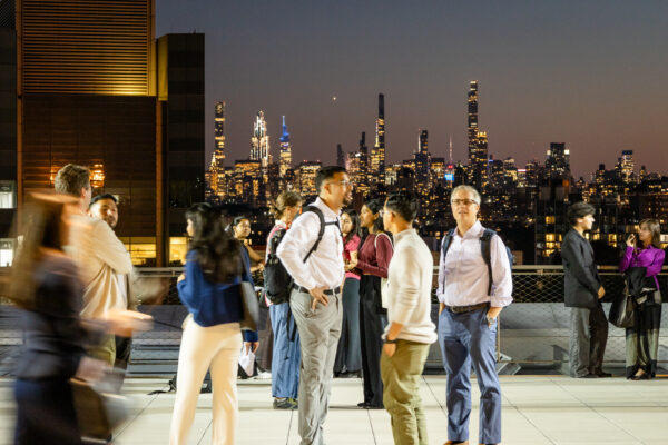 A group of diverse people are mingling and conversing on an outdoor terrace or rooftop at night. The brightly lit Manhattan skyline, including several tall skyscrapers, forms a dramatic backdrop to the networking event. Some individuals in the foreground are slightly blurred due to movement.