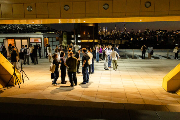 A large group of attendees is mingling and standing in small clusters on a wide, tiled rooftop terrace at night. The view is framed by a low, golden-yellow overhang structure. In the background, the lit-up city skyline is visible against the dark night sky.