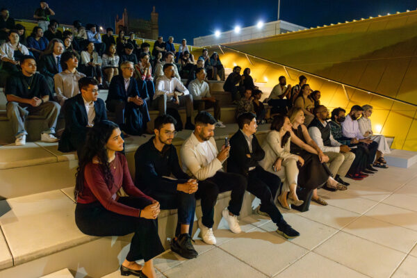 A large group of diverse attendees is seated on stepped, tiered seating outdoors at night, resembling an amphitheater. Everyone is facing toward the right, listening to or watching a presentation. A yellow-green sloped architectural feature is visible on the right, and a dark building with a pointed roof is visible in the background.