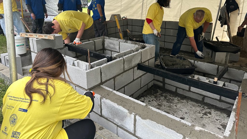 A group of architecture students and professional masons, all wearing bright yellow shirts, collaborate on building a concrete masonry unit (CMU) structure. The students are actively laying blocks and mortar, guided by the experienced tradespeople. The structure appears to be a planter or a small wall, and the setting is outdoors under a tent, suggesting a hands-on workshop or competition.