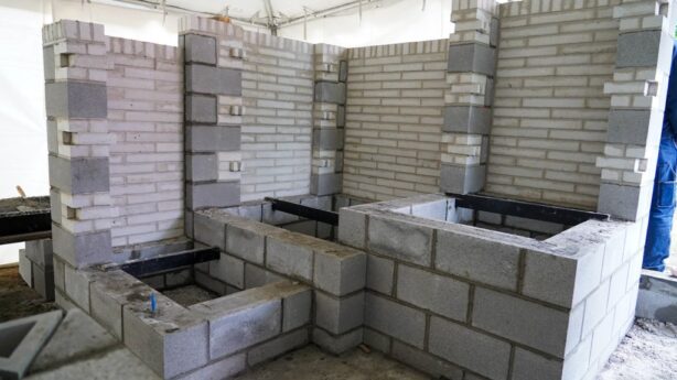 A full-scale mock-up of a wall and planter system is constructed from a combination of gray concrete masonry units (CMU) and light-colored bricks. The structure shows different masonry techniques and patterns, including both stacked blocks and staggered brickwork, highlighting the various ways the materials can be used together. The structure is built on a custom platform under a large tent.