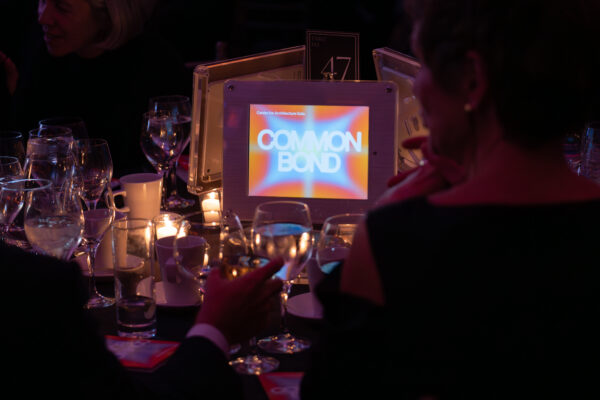 A close-up, dark photo of a round dinner table setting at the Center for Architecture Gala. The center of the table features a small digital screen displaying the event logo: the words 