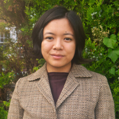 A headshot of Justine Flora, a young woman with dark hair and a nose ring, standing outside and looking directly at the camera. She is wearing a brown, speckled tweed blazer over a darker shirt, and is framed against a background of lush, green shrubbery.