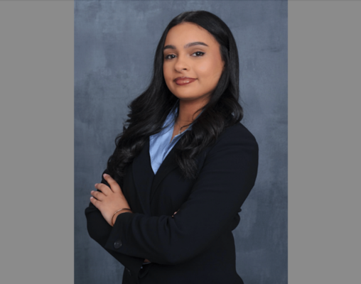 A professional headshot of a young woman with long, wavy dark hair and a warm, confident expression. She is posed at a slight angle with her arms crossed, looking toward the camera. She is wearing a dark navy blue or black blazer over a light blue collared shirt. The background is a textured, mottled grey-blue studio backdrop. The lighting is soft and even, highlighting her polished makeup and jewelry, which includes a gold bracelet and a delicate necklace.