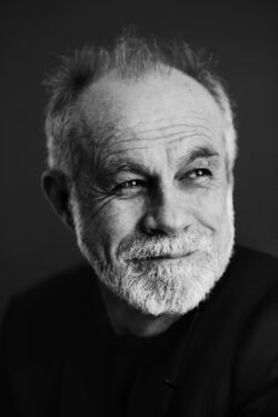 A black-and-white portrait of Carlos Moreno, a man with a short white beard and graying hair. He is looking slightly away from the camera with a thoughtful expression and a faint smile. The lighting creates high contrast, emphasizing his facial features against a dark, minimalist background.