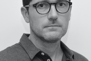 A black-and-white headshot of Martín Cobas, a man with short dark hair and dark-rimmed glasses, wearing a polo shirt and looking directly at the camera with a neutral expression.
