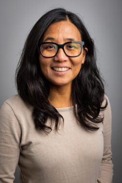 A woman with long black hair, wearing black glasses and a tan sweater, smiles for a headshot against a grey background.
