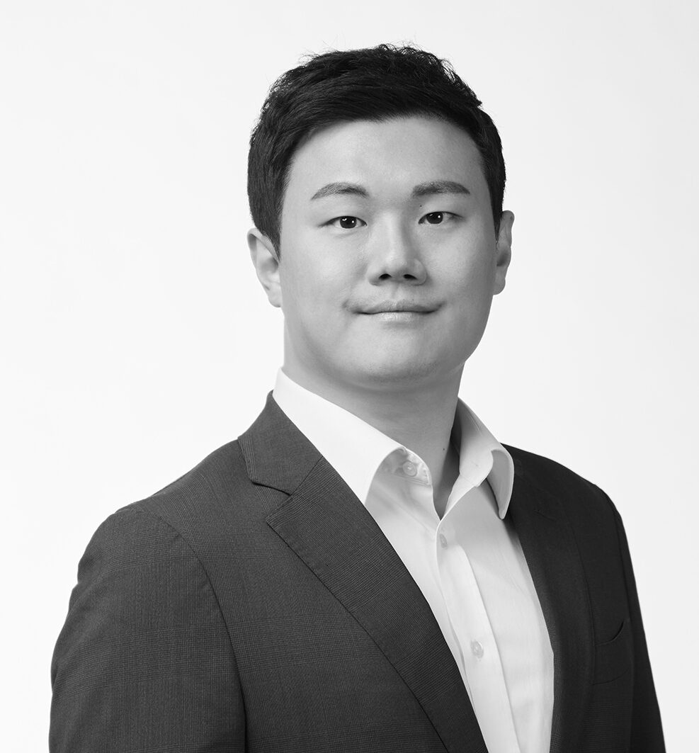 A black and white, waist-up portrait of a man with short, dark hair, wearing a dark suit jacket over a light-colored, button-down shirt. He is looking directly at the camera with a slight, pleasant smile against a plain, light-colored background.