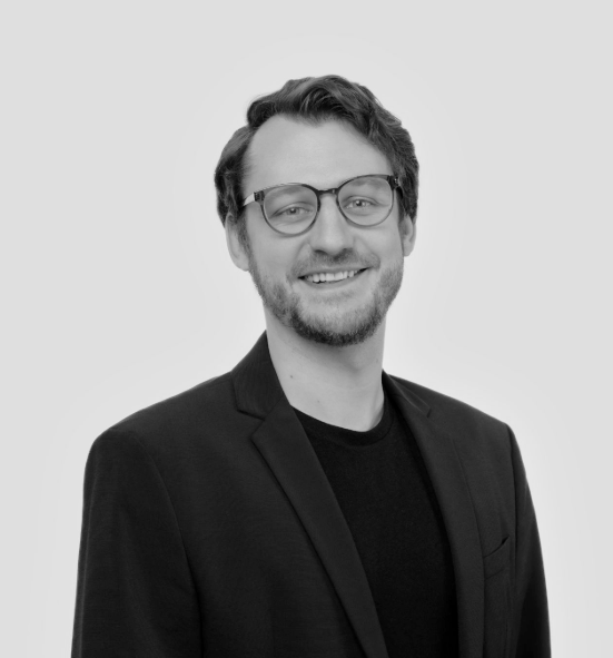 A black-and-white headshot of a smiling man with short, wavy hair, a light beard, and glasses. He is wearing a dark t-shirt under a dark blazer and is positioned against a solid light gray background.