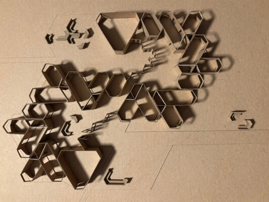An overhead photograph of a conceptual architectural model made from brown cardboard. The model consists of numerous interlocking and individual geometric shapes, primarily hexagons and elongated polygons, arranged in a sprawling, organic pattern across a flat cardboard base. Some shapes are grouped closely together to form dense clusters, while others are more isolated. Faint geometric lines are etched into the base board, suggesting a site plan or layout. Dramatic lighting from the side casts long, deep shadows, emphasizing the three-dimensional forms and the varying heights of the cardboard walls.
