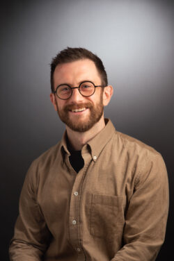 A headshot of a smiling man with a beard and round, black-rimmed glasses, wearing a tan corduroy button-down shirt over a black t-shirt against a dark grey gradient background.